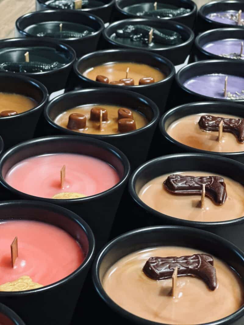 An assortment of scented candles with varying colors and some with embedded shapes, freshly made in black tins and waiting to solidify.