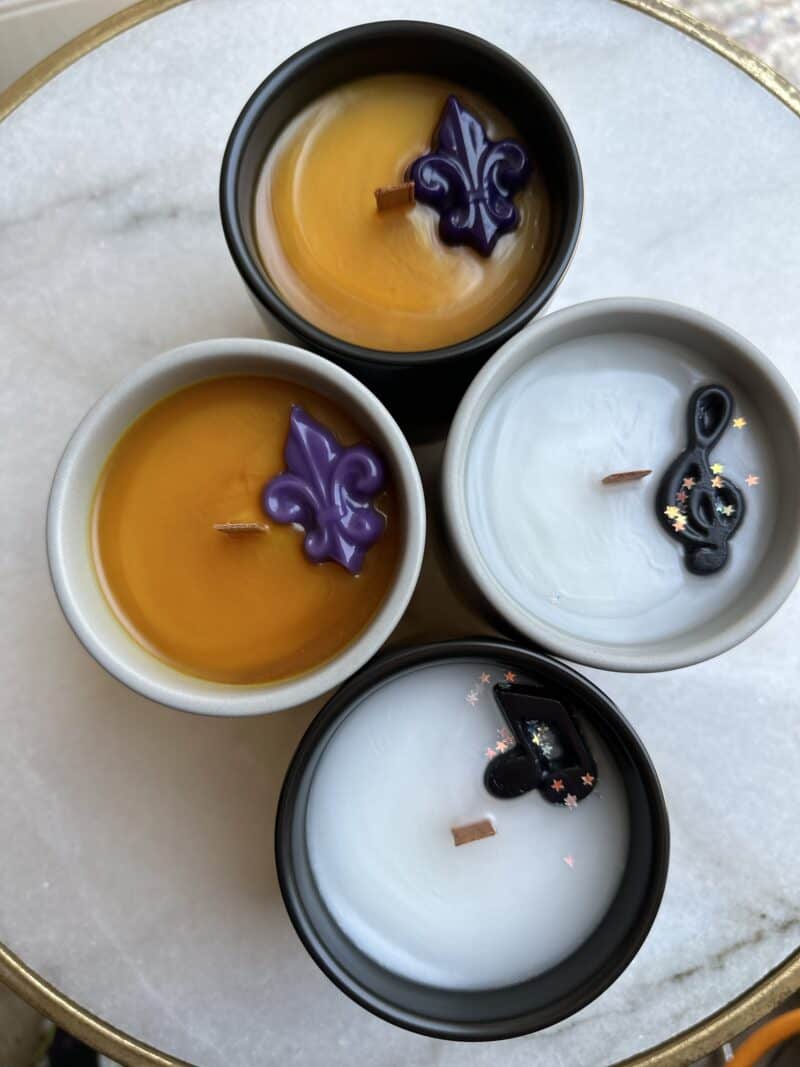 Four candles in containers placed on a surface, with three having white wax and decorative elements on top, and one with a yellowish-orange wax.