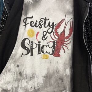 The Feisty & Spicy Crawfish Shirt is a grey tee featuring "Feisty & Spicy" text with images of a red crawfish, lemon slices, and corn on the cob, shown partially covered by a dark jacket.